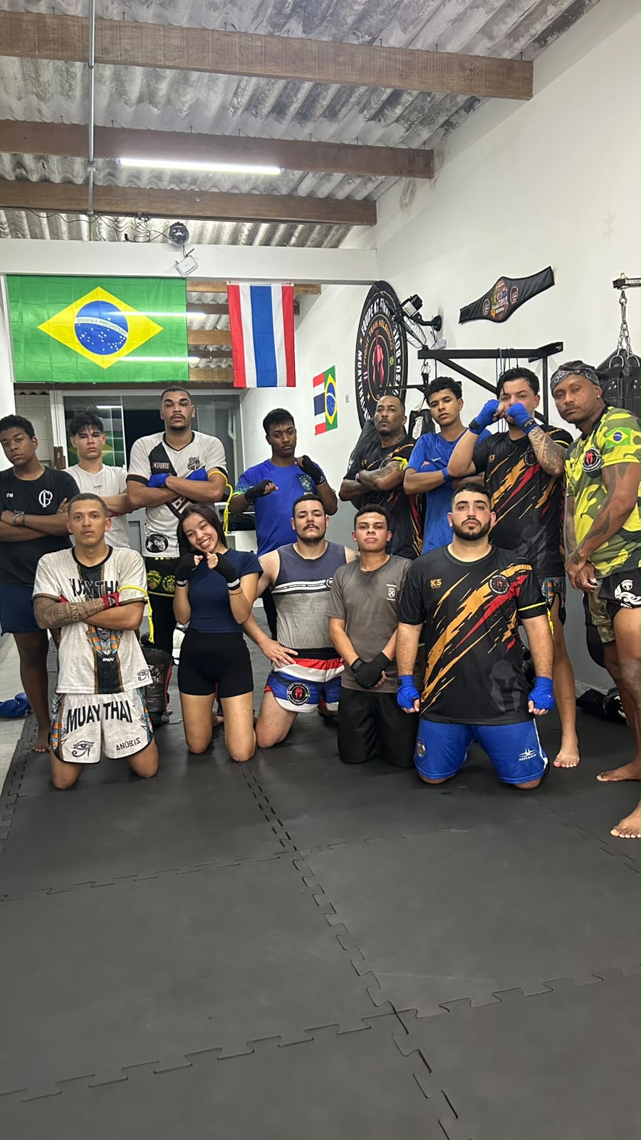 Turma cheia reunida no tatame do CT sob as bandeiras do Brasil e da Tailândia